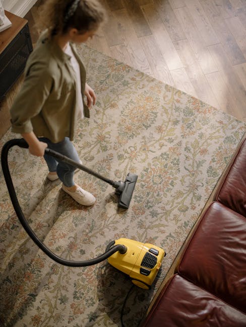 A professional in full protective cleaning attire, including a white jumpsuit, face mask, and blue gloves, is performing deep cleaning on a beige plush carpet using a professional steam cleaning machi