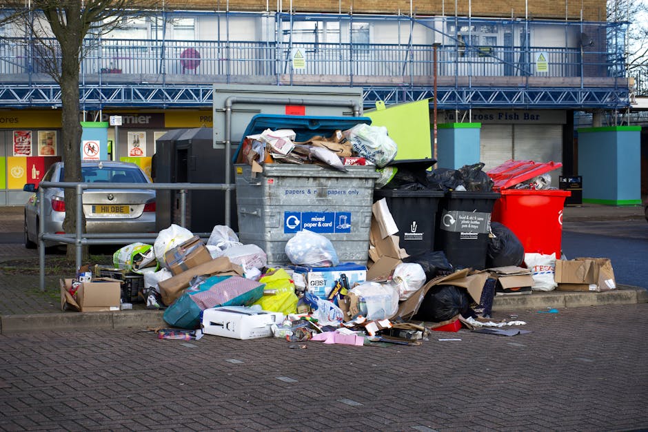A large pile of mixed household and commercial rubbish including cardboard boxes, plastic bags, paper, and discarded packaging on a paved sidewalk near a commercial building with storefronts in the ba