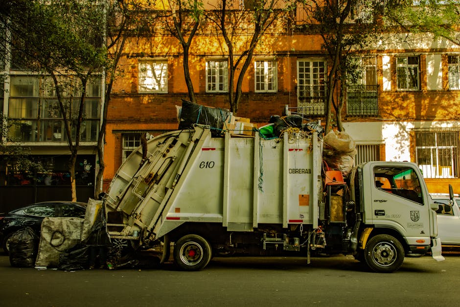 A large pile of mixed household and commercial rubbish including cardboard boxes, plastic bags, paper, and discarded packaging on a paved sidewalk near a commercial building with storefronts in the ba