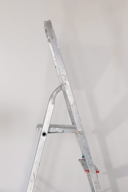 A silver aluminum step ladder with four steps, positioned against a plain, light-colored wall in a room with bright, even lighting. The ladder's surface shows minor signs of wear and is free of clutter or dust, indicating recent or ongoing cleaning activities. The background wall appears clean and smooth, with no visible stains or marks, suggesting a well-maintained environment. The presence of the ladder may imply preparations for cleaning or maintenance work, consistent with surface cleaning and deep cleaning processes by Harringay Carpet Cleaning. No other objects or furniture are visible in the image, highlighting the focus on the ladder and the clean, tidy space.