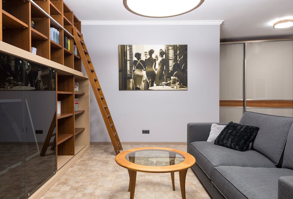 A modern living room featuring a grey fabric sofa with black and white cushions, a wooden coffee table with a glass top, and a large wooden bookshelf on the left filled with books and decorative items. A wooden ladder leans against the bookshelf, leading up to additional storage. On the white wall behind the sofa, there is a black-and-white photograph of women in vintage dresses near a window with shutters. The room is well-lit with ceiling-mounted circular light fixtures, the floor is covered with light-colored wooden or laminate flooring, and the space appears clean and tidy, reflecting professional domestic cleaning services by Harringay Carpet Cleaning.