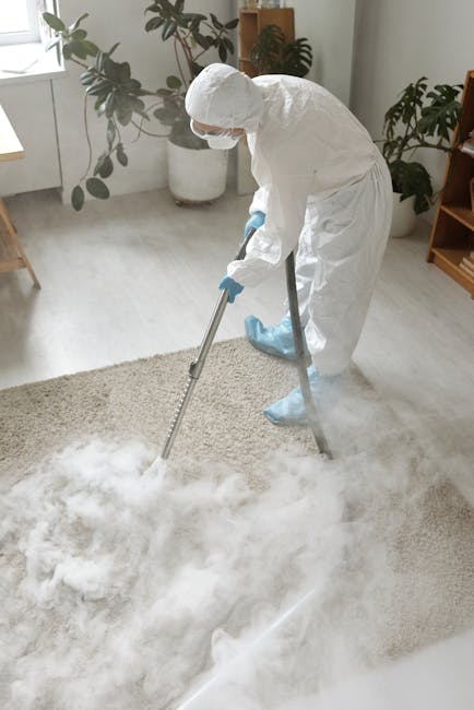 A person kneeling on a patterned carpet in a domestic setting, using a yellow and black vacuum cleaner with a detachable handheld unit for surface cleaning. The individual is wearing a beige jacket an