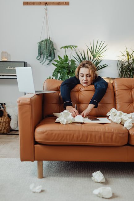 A person dressed in full protective PPE, including a white coverall suit, mask, and gloves, is performing surface cleaning on a black leather sofa in a well-lit living room. The sofa is situated near